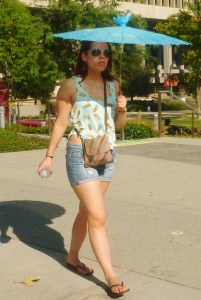 20 something woman in shorts and and colorful blouse holds a parasol while walking down hill street 