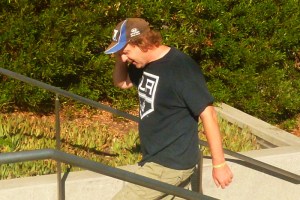 male 30 something with LA Kings shrit and baseball cap walks up the steps in park with head phones on to his smart phone