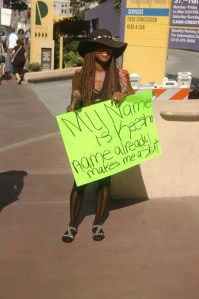 Africian aAmercian 30 something woman in sexy black dress and hat hold poster at Slutwalk